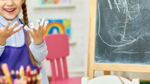 Portrait of cute little girl enjoying art and craft lesson in development school and smiling happily looking at hands covered in paint